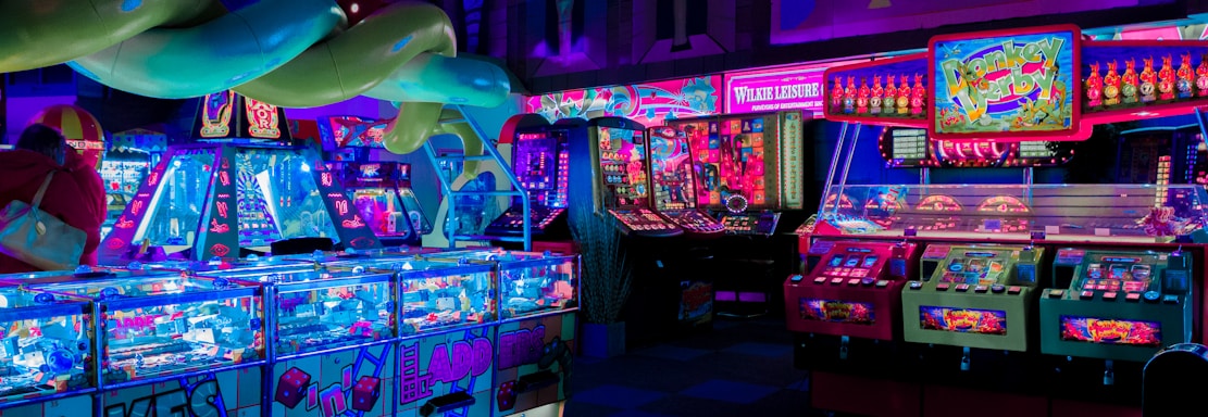 An indoor arcade filled with vibrant gaming machines lit with neon blue and pink lights. The scene includes various arcade games with flashing screens and colorful displays, and a large overhead structure resembling a green snake. The background shows illuminated signs and themed decor, contributing to an immersive entertainment atmosphere.