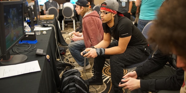 Several people are sitting in a room with gaming consoles and monitors, focusing intently on their screens. The setting appears to be a casual gaming event, with participants holding controllers while playing video games. The room has a casual atmosphere with chairs and tables set up for gaming.