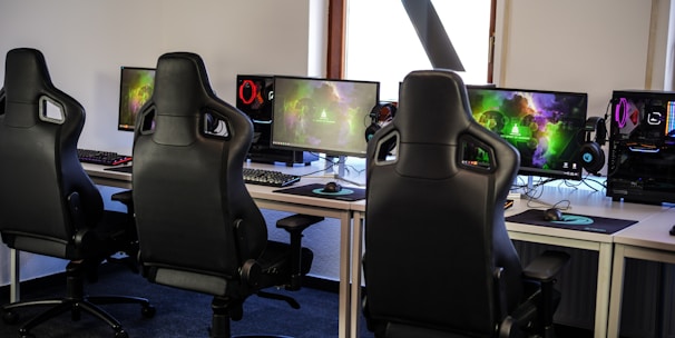 A row of gaming stations equipped with large monitors, gaming chairs, keyboards, and headphones. The setup is organized and ready for gaming, featuring a colorful screen display. The room has a modern and tech-savvy ambiance.