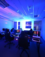 A modern gaming setup with multiple high-backed gaming chairs and monitors arranged in a room with a cool blue lighting. The computers have illuminated keyboards and glowing screens, creating a high-tech ambiance.