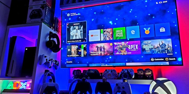 A modern gaming setup featuring a large wall-mounted screen displaying a gaming console interface. The room is decorated with gaming-themed items such as controllers and collectibles. The ambient lighting includes shades of blue and red, creating a vibrant atmosphere.