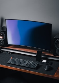 A modern gaming and streaming setup with a curved monitor placed centrally on a wooden desk. Surrounding the monitor are powerful speakers and a high-end PC tower with glowing fans. Various peripherals, including a mechanical keyboard, ergonomic mouse, and audio mixer, are neatly arranged. Overhead, a professional microphone and stand are ready for use, possibly for gaming commentary or podcasting.