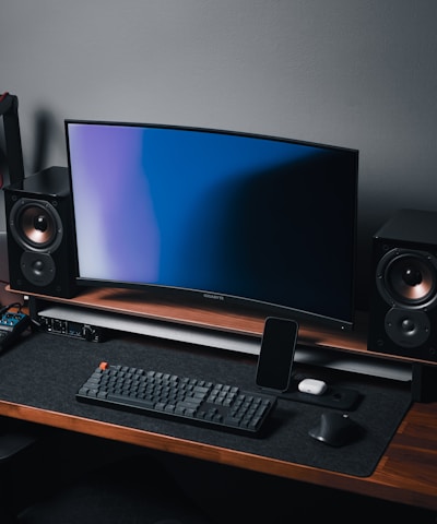 A modern gaming and streaming setup with a curved monitor placed centrally on a wooden desk. Surrounding the monitor are powerful speakers and a high-end PC tower with glowing fans. Various peripherals, including a mechanical keyboard, ergonomic mouse, and audio mixer, are neatly arranged. Overhead, a professional microphone and stand are ready for use, possibly for gaming commentary or podcasting.
