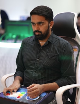 A person is seated in a chair, focused intently on something, with hands positioned on a gaming console or arcade controller. The setup appears to be indoors, with visible screens and lighting in the background, suggesting an electronic gaming environment.